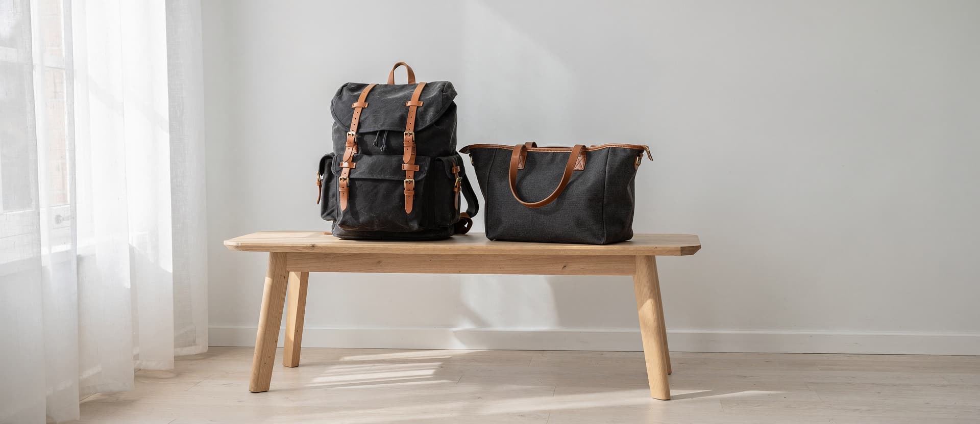 A waxed-canvas backpack and a canvas tote on an oak bench by a window